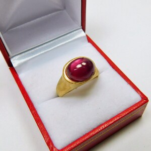 Puede incluir: Un anillo de oro con una gran piedra preciosa ovalada de color rojo oscuro engastada en un engaste de oro. El anillo está en una caja de anillo de terciopelo rojo.