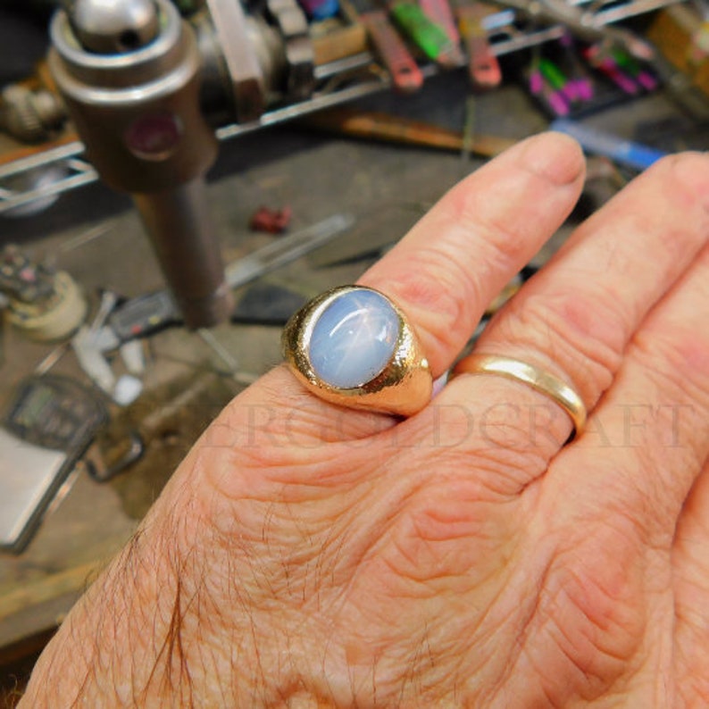 Puede incluir: Un anillo de oro con una gran piedra preciosa azul de forma ovalada engastada en el centro. La piedra preciosa tiene un patr&oacute;n en forma de estrella.