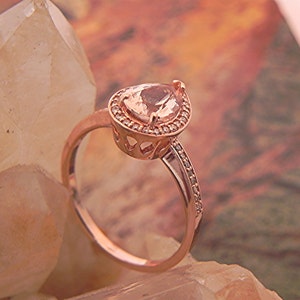 May include: A rose gold ring with a pear-shaped gemstone set in a halo of smaller stones. The ring is on a background of white and brown crystals.