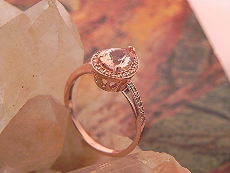May include: A rose gold ring with a pear-shaped gemstone set in a halo of smaller stones. The ring is on a background of white and brown crystals.