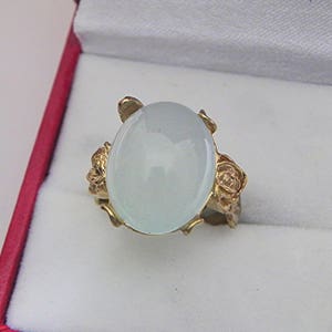 Puede incluir: Un anillo de oro con una gran piedra blanca ovalada y dos acentos en forma de rosa a cada lado.