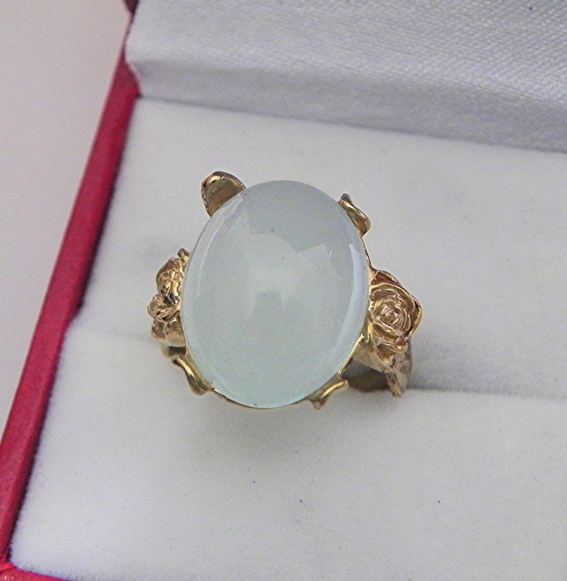 Puede incluir: Un anillo de oro con una gran piedra blanca ovalada y dos acentos en forma de rosa a cada lado.