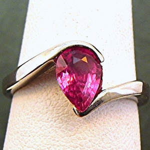 May include: A pear-shaped pink gemstone ring set in a silver band with a curved design.
