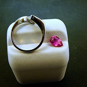 May include: A silver ring with a pear-shaped pink gemstone on a white display stand.