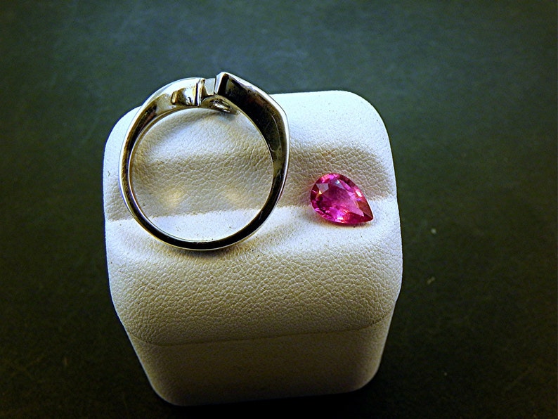 May include: A silver ring with a pear-shaped pink gemstone on a white display stand.