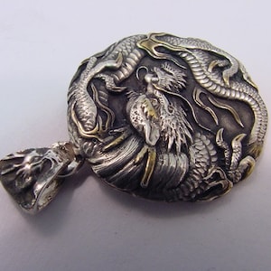 May include: A silver and gold pendant featuring a detailed dragon design. The pendant is round and has a textured surface. The dragon is depicted in a curled position with its tail wrapped around its body. The pendant has a loop at the top for attaching to a chain.