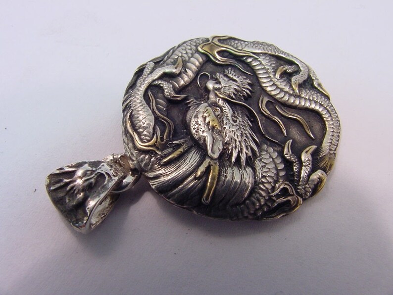 May include: A silver and gold pendant featuring a detailed dragon design. The pendant is round and has a textured surface. The dragon is depicted in a curled position with its tail wrapped around its body. The pendant has a loop at the top for attaching to a chain.