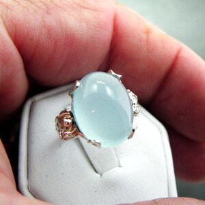 Puede incluir: Un anillo de plata con una gran piedra preciosa ovalada de color azul claro. El anillo tiene intrincados detalles de plata alrededor de la piedra preciosa.