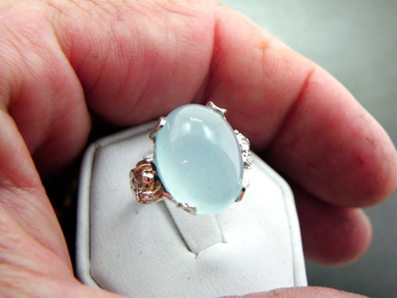 Puede incluir: Un anillo de plata con una gran piedra preciosa ovalada de color azul claro. El anillo tiene intrincados detalles de plata alrededor de la piedra preciosa.