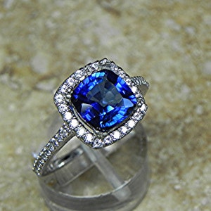 Puede incluir: Un anillo de plata con un gran zafiro azul de talla cojín rodeado por un halo de diamantes más pequeños.