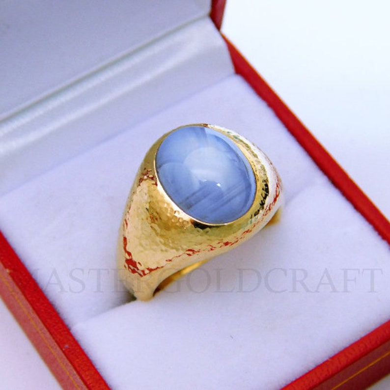 Puede incluir: Un anillo de oro con una gran piedra preciosa azul de forma ovalada engastada en el centro. El anillo est&aacute; en una caja de terciopelo rojo.