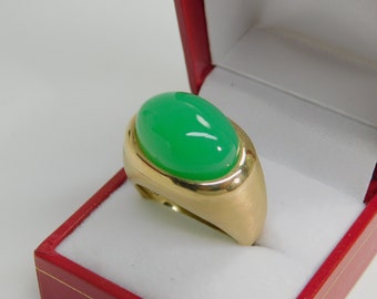 May include: A gold ring with a large, oval-shaped green gemstone set in a smooth, rounded bezel. The ring is displayed in a red velvet ring box.