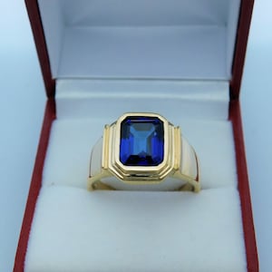 May include: A gold ring with a blue gemstone set in a square shape. The ring is in a red and white box.
