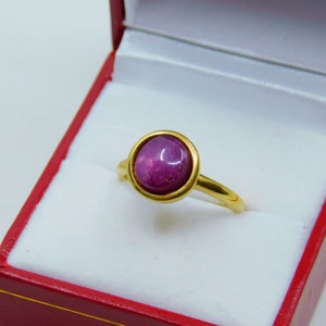 May include: A gold ring with a round, purple gemstone set in a bezel setting. The ring is in a red jewellery box.
