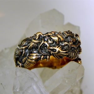 Puede incluir: Un anillo de tono dorado con un diseño de dragón detallado. La cabeza del dragón es prominente, con escamas intrincadas y una expresión feroz.
