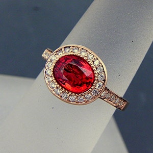 May include: A rose gold ring with a large oval red gemstone surrounded by a halo of smaller white gemstones.