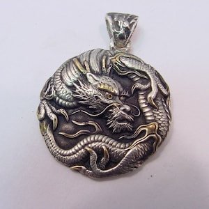 Puede incluir: Un colgante de dragón de plata y oro con detalles intrincados. El dragón está enroscado sobre sí mismo, con la cabeza mirando hacia adelante. El colgante tiene forma circular y tiene un bucle en la parte superior para sujetarlo a una cadena.