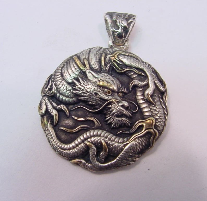 May include: A silver and gold dragon pendant with intricate details. The dragon is curled around itself, with its head facing forward. The pendant is a circular shape and has a loop at the top for attaching to a chain.