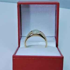 K&ouml;nnte beinhalten: Ein goldener Ring mit einem hellgr&uuml;nen ovalen Edelstein, der in einer Zargenfassung gefasst ist. Der Ring befindet sich in einer roten Samt-Ringbox.