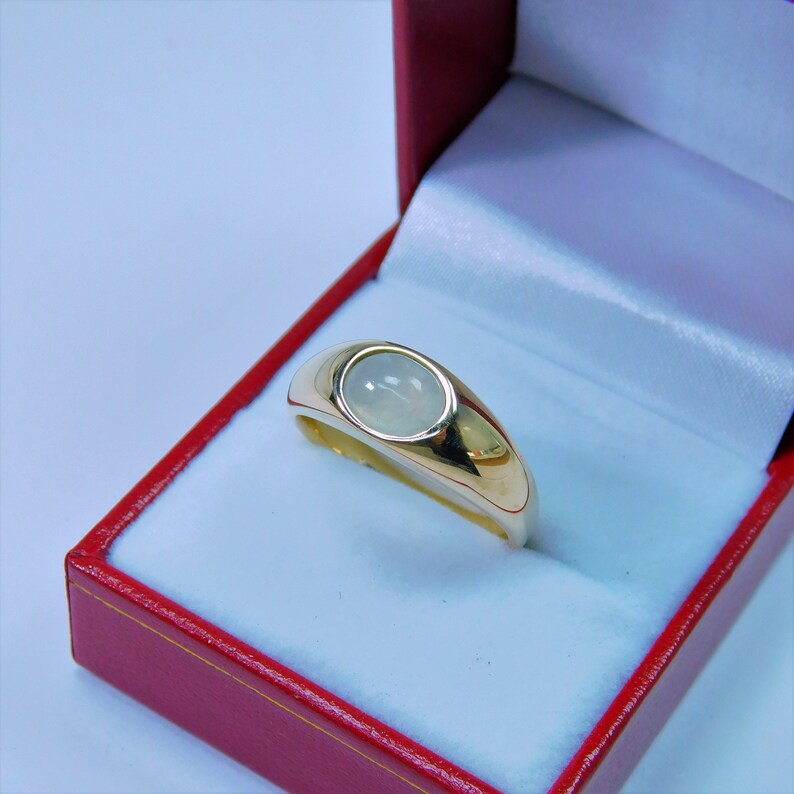 K&ouml;nnte beinhalten: Ein goldener Ring mit einem wei&szlig;en ovalen Edelstein, der in einer Zargenfassung gefasst ist. Der Ring befindet sich in einer roten Samtringschatulle.