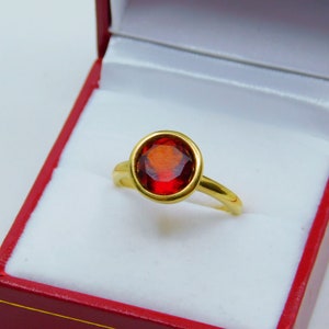 Puede incluir: Un anillo de oro con una piedra preciosa roja en el centro. El anillo est&aacute; en una caja de terciopelo rojo.