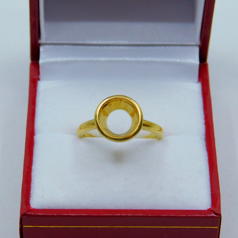 May include: A gold ring setting with a round bezel, ready to be set with a gemstone. The ring is displayed in a red velvet box.