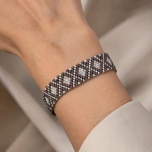 Puede incluir: Una pulsera de cuentas tejidas con un patrón geométrico repetitivo de diamantes en cuentas de vidrio de color plata metálico, gris oscuro y blanco. La pulsera tiene una estructura de banda ancha y plana con una textura de rejilla uniforme.
