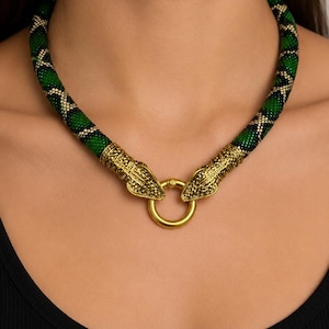 May include: A beaded necklace featuring a green and black snake-like pattern with gold-toned metal ends shaped like animal heads. The two heads meet at the center, connected by a circular gold-toned ring clasp that secures the necklace.