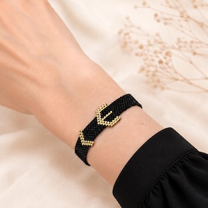 May include: A black woven bracelet featuring a gold-toned buckle-style clasp. The bracelet is constructed from small, uniform beads arranged in a tight, grid-like pattern, with the metallic gold buckle element serving as the central focal point.