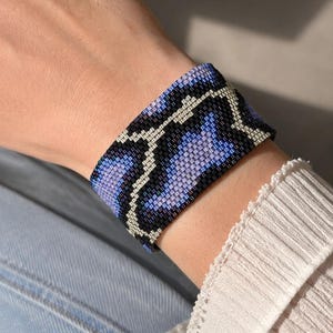 May include: A wide cuff bracelet made of small glass beads arranged in a geometric pattern. The design features alternating sections of black, light purple, and metallic silver beads, creating a repeating wave-like motif across the entire surface.