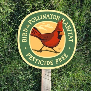 May include: A wooden garden stake with a circular sign. The sign features a red cardinal illustration and text that reads "Bird & Pollinator Habitat" and "Pesticide Free" in a green and yellow design. The stake is set in green grass.