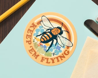 May include: A round sticker with a bee flying over a field of flowers. The sticker has the text "Keep 'em Flying" in a retro font.