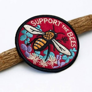 May include: A red embroidered patch with a bee and flowers. The text "Support the Bees" is embroidered in white on the patch.