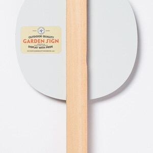 May include: A light gray, oval-shaped garden sign with a natural wood handle. The sign features text that reads "Outdoor Quality Garden Sign" and "Display with Pride" on a cream-colored label. The handle is made of light-colored wood.