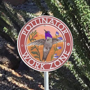 May include: A round sign with the words "POLLINATOR WORK ZONE" in white lettering. The sign features a hummingbird with outstretched wings, purple head, and gray body, set against a landscape of orange and blue hues. The sign is mounted on a metal pole.