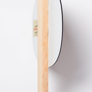 May include: A white, round garden display sign with a black border, attached to a natural wood stake. The sign has a small label with text. The sign is designed for outdoor use.