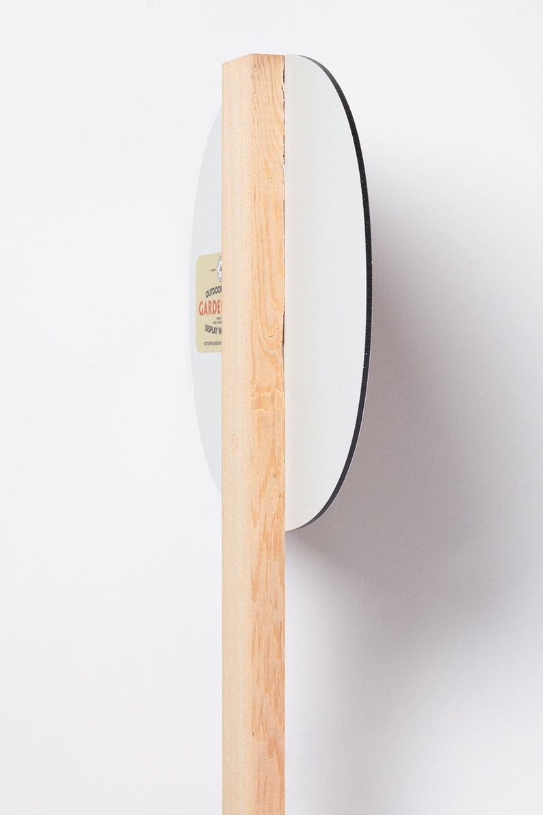 May include: A white, round garden display sign with a black border, attached to a natural wood stake. The sign has a small label with text. The sign is designed for outdoor use.