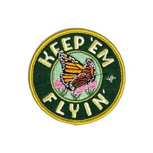 Puede incluir: Un parche bordado verde y dorado con una mariposa y el texto "Keep 'em Flyin'"