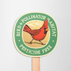May include: A round sign with a green border and the words "Bird & Pollinator Habitat Pesticide Free." The sign features a red cardinal bird illustration. The sign is attached to a wooden stake.