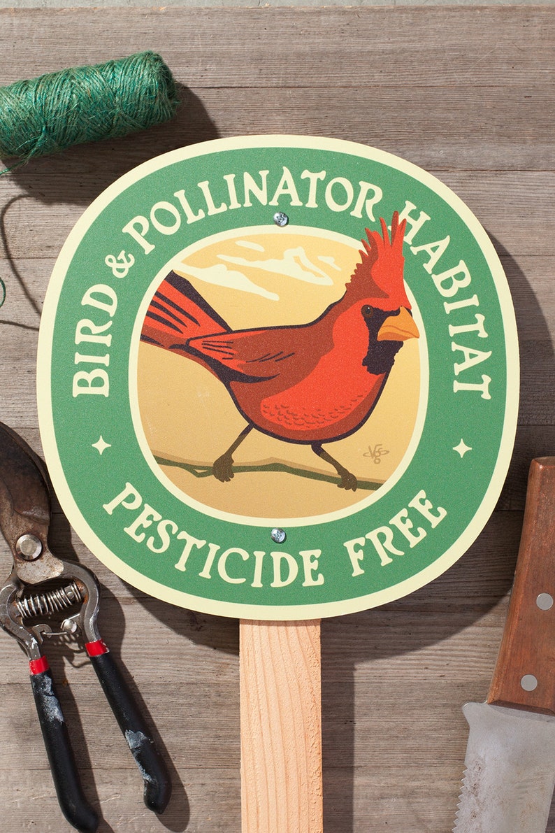 May include: A round green sign with the text "BIRD & POLLINATOR HABITAT PESTICIDE FREE" surrounds a red cardinal illustration. The sign is mounted on a wooden stake, with gardening tools and twine in the background.