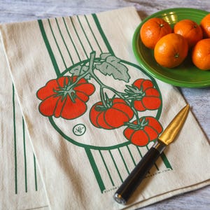 May include: A white tea towel with green stripes and a red tomato vine illustration. The towel has the text "VICTORIA GROW COM" printed on it. A black handled knife is lying on the towel.