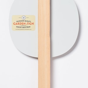 May include: A white round garden sign with a wooden stake. The sign has a sticker that says "Outdoor Quality Garden Sign Printed in Portland, Oregon Display with Pride VictoryGardenofTomorrow.com".