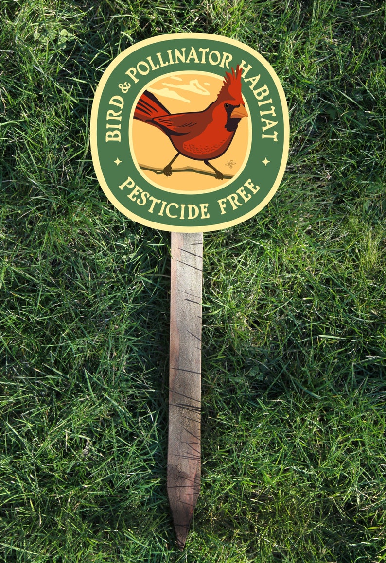 May include: A wooden garden stake with a circular sign. The sign features a red cardinal illustration and text that reads "Bird & Pollinator Habitat" and "Pesticide Free" in a green and yellow design. The stake is set in green grass.