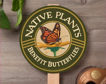 May include: A round green sign with the text "NATIVE PLANTS BENEFIT BUTTERFLIES" in a gold circle with a monarch butterfly on a yellow flower. The sign is attached to a wooden stake.