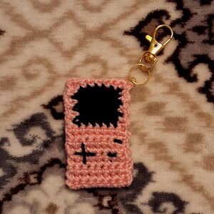 May include: A pink crocheted keychain shaped like a handheld video game console. The keychain has a black screen and buttons.