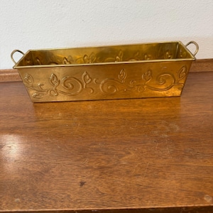 May include: A rectangular brass planter featuring a polished metallic finish and two small side handles. The exterior walls are decorated with an embossed vine and leaf pattern that wraps around the front and sides of the elongated container.