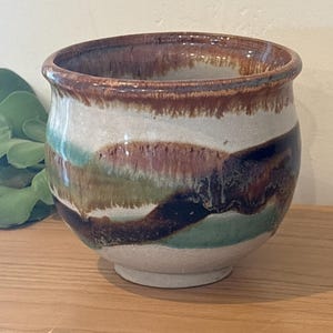 May include: A rounded ceramic bowl with a speckled off-white base and a wide, flared rim. The exterior features horizontal, flowing bands of glaze in shades of deep brown, forest green, and teal, creating a layered, organic, and multi-colored appearance.