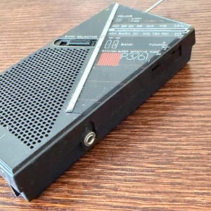 May include: Black vintage portable radio with a silver antenna and a speaker grill. The radio has a band selector, volume control, and tuning dial. The text "P3761" is visible on the front. The radio is designed to receive FM and AM radio frequencies.