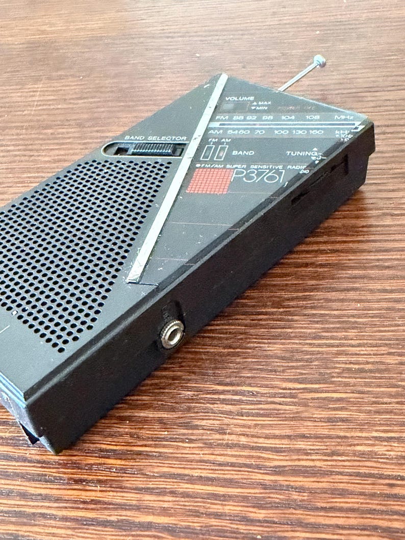 May include: Black vintage portable radio with a silver antenna and a speaker grill. The radio has a band selector, volume control, and tuning dial. The text "P3761" is visible on the front. The radio is designed to receive FM and AM radio frequencies.
