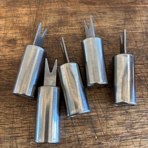 May include: Five stainless steel cheese markers, each with a cylindrical body and a forked top. They are displayed on a wooden surface, reflecting light.